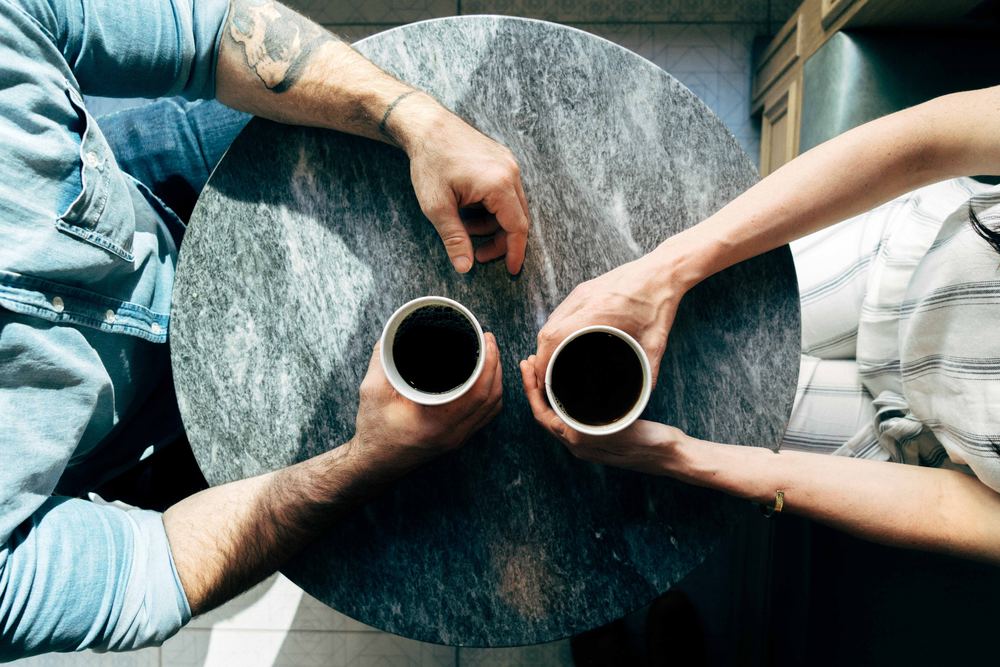 An overhead photo of two people sitting at a table with coffee. Only the coffee and their hands are visible.