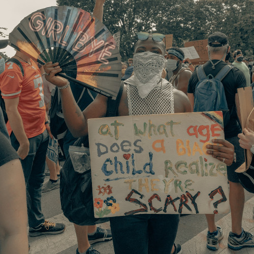 A man at a Black Lives Matter protest holding up a sign that says, "At what age does a black child realize they're scary?"
